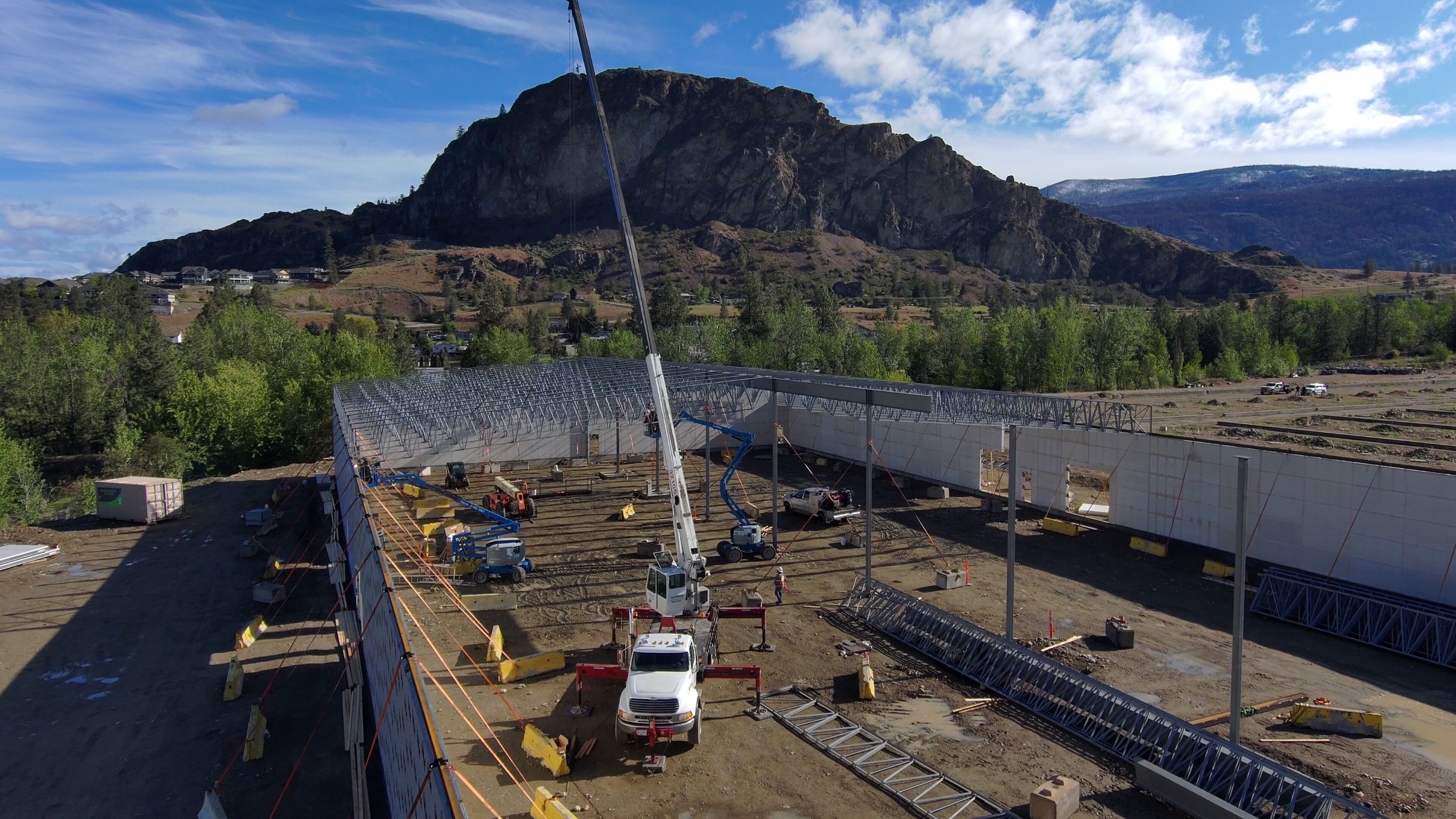 An EcoTech prefabricated facility under construction, steel roof structure being lifted into place against a mountain backdrop